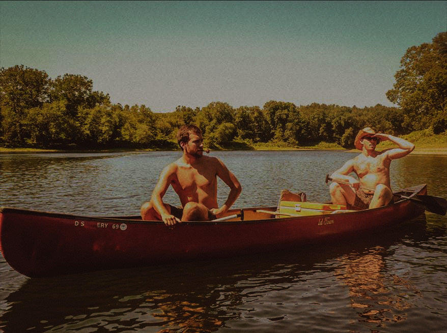 Wild Hickory founders sitting in a red canoe on a placid lake, peering into the distance while smoking a pipe.